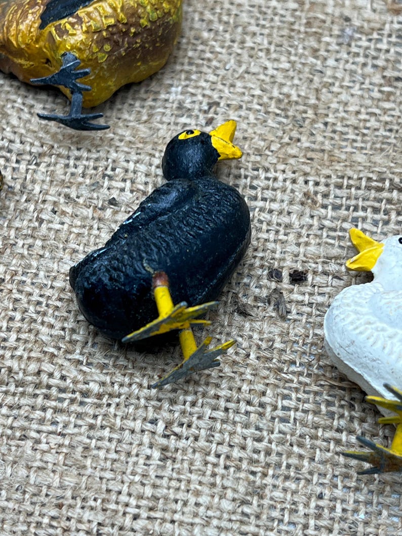 K&ouml;nnte beinhalten: Eine Sammlung kleiner, bemalter Vogelfiguren. Ein schwarzer Vogel mit gelbem Schnabel und F&uuml;&szlig;en steht im Fokus. Weitere V&ouml;gel sind zu sehen, darunter ein gelber und ein wei&szlig;er Vogel. Die Figuren befinden sich auf einer strukturierten, beigen Oberfl&auml;che.