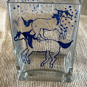 May include: A clear glass square vase with a blue and white painted design of three horses running. The background is a light blue with white dots.