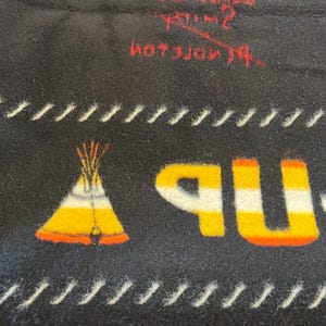 Puede incluir: Manta negra con un gr&aacute;fico de tipi nativo americano en amarillo, naranja y blanco. La palabra "UP" est&aacute; en amarillo y naranja. L&iacute;neas cosidas blancas bordean el dise&ntilde;o. Texto rojo visible en la parte superior.