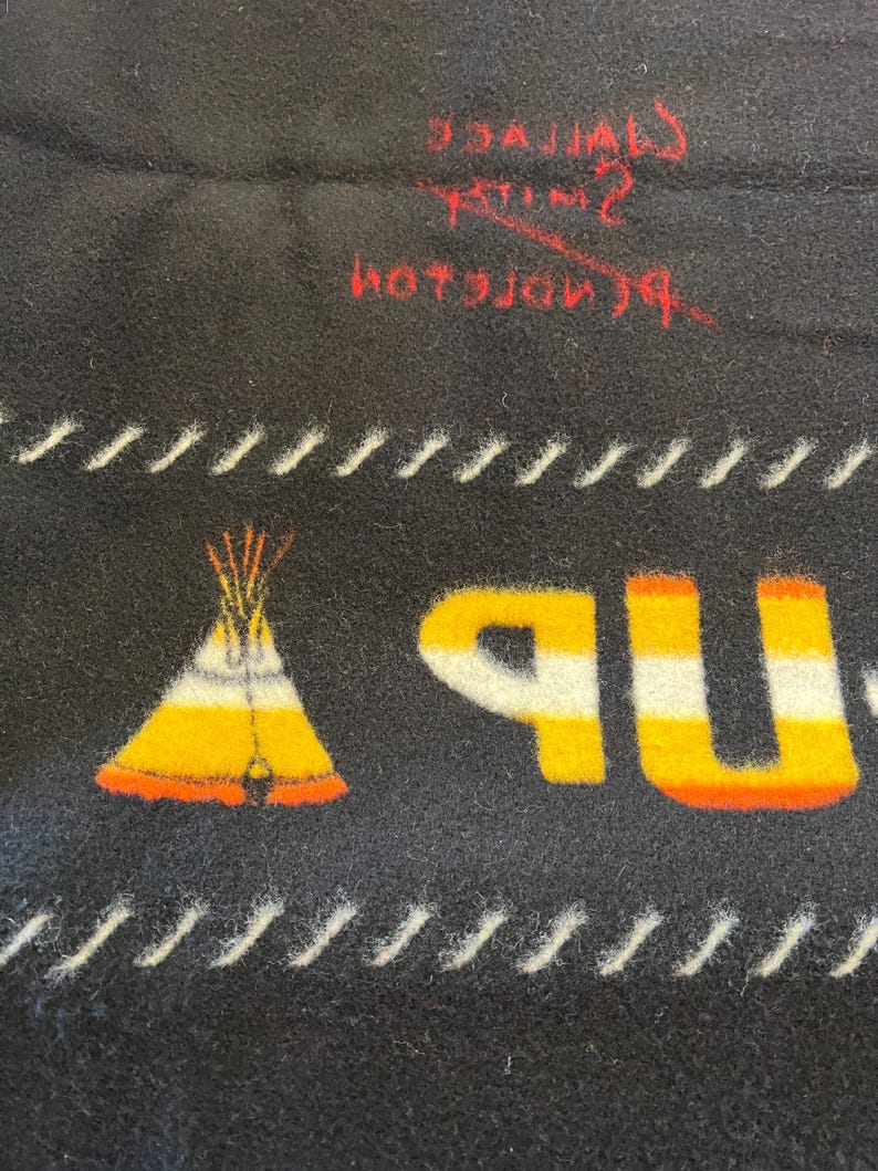 Puede incluir: Manta negra con un gr&aacute;fico de tipi nativo americano en amarillo, naranja y blanco. La palabra "UP" est&aacute; en amarillo y naranja. L&iacute;neas cosidas blancas bordean el dise&ntilde;o. Texto rojo visible en la parte superior.