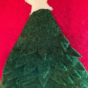 May include: A handmade Christmas tree decoration. The tree is green with a white star on top, set against a red background. The tree is made of felt and has a textured appearance, suggesting a handcrafted item.