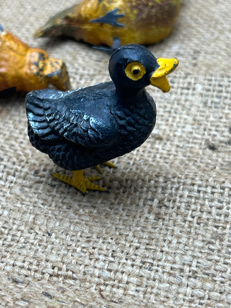 K&ouml;nnte beinhalten: Eine kleine, schwarze Vogelfigur mit gelbem Schnabel und F&uuml;&szlig;en. Der Vogel hat gelbe Augen und steht auf einer strukturierten Oberfl&auml;che. Weitere Vogelfiguren sind im Hintergrund zu sehen.