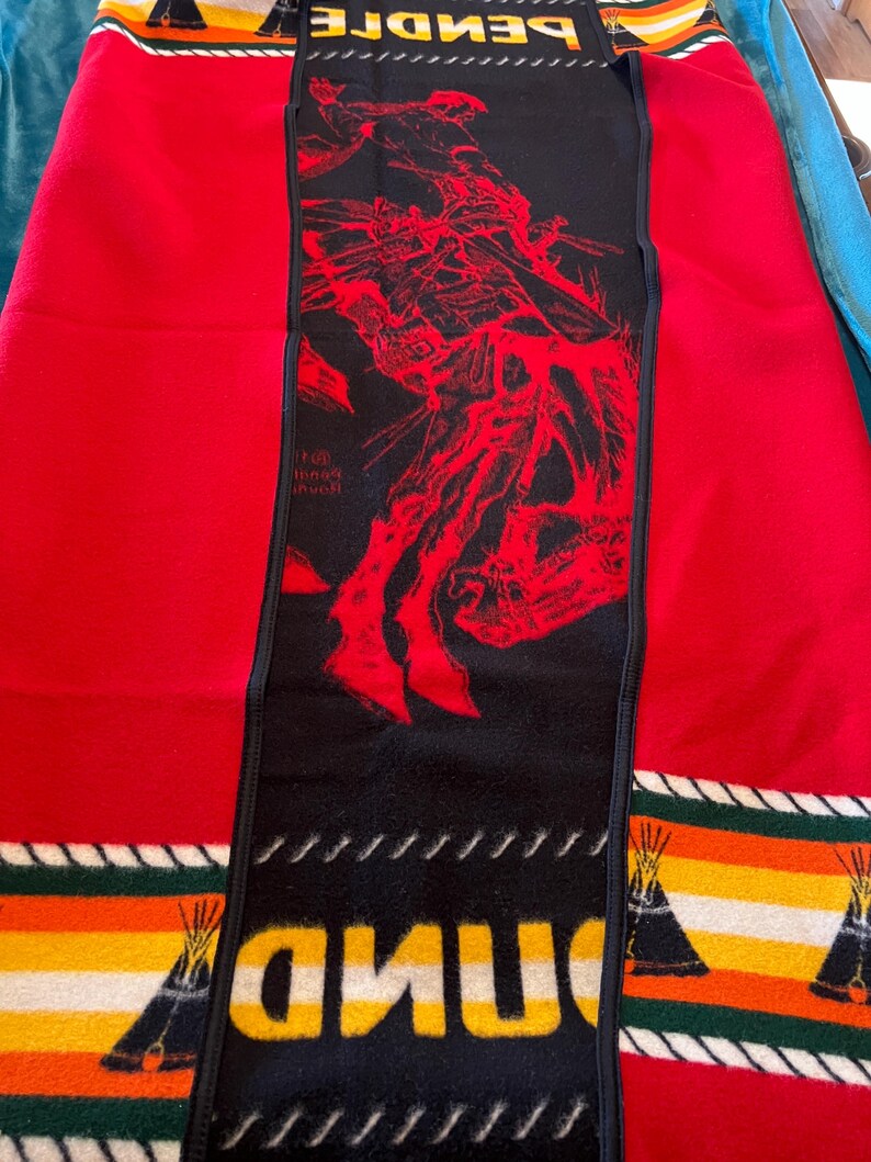 Puede incluir: Una manta roja y negra con un dise&ntilde;o nativo americano. La manta presenta un panel central negro con una ilustraci&oacute;n roja y la palabra "PENDLE" en amarillo. Los bordes tienen patrones de rayas con dise&ntilde;os de tipi.