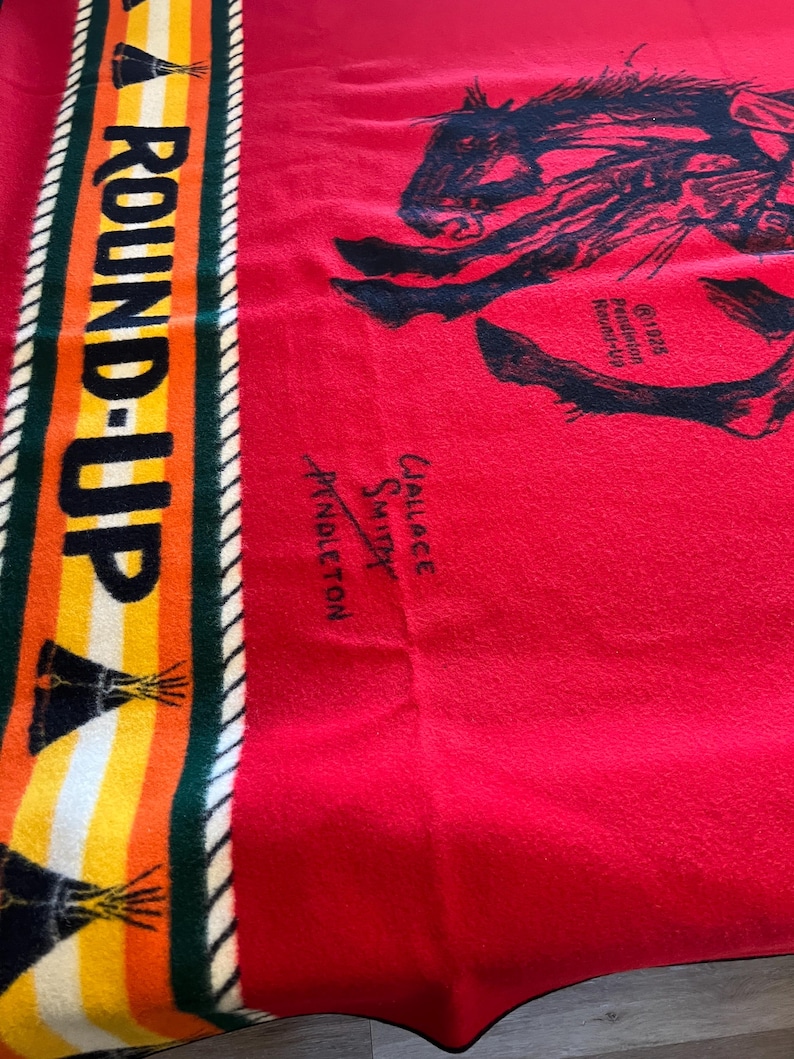 Puede incluir: Una manta roja Pendleton Round-Up con una ilustraci&oacute;n de caballo negro. La manta tiene un borde a rayas con las palabras "ROUND-UP" en amarillo y naranja. La manta tambi&eacute;n tiene el texto "1926 Pendleton Round-Up" y una firma.
