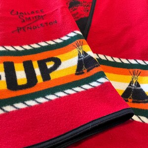 Puede incluir: Manta de lana roja con borde negro y dise&ntilde;o de rayas coloridas. La manta presenta la palabra "UP" en letras amarillas grandes, un gr&aacute;fico de tipi y el texto "Wallace Smith Pendleton".