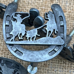 May include: Antique silver-colored horseshoe-shaped cabinet knobs. Each knob features a cutout design of a cowboy riding a horse, with a second horse and rider. The knobs have a screw for easy installation.