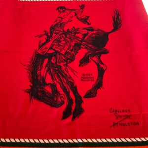 Puede incluir: Una manta roja de Pendleton con una ilustraci&oacute;n en negro de un vaquero montando un bronco. La manta tiene un borde con las palabras "PENDLETON" y "ROUND-UP" y un dise&ntilde;o de tipi nativo americano. La manta est&aacute; firmada por Wallace Smith.