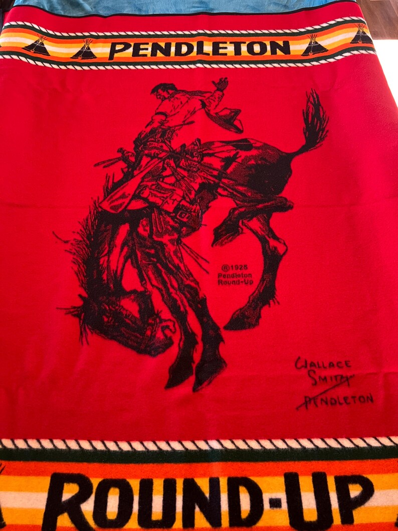 Puede incluir: Una manta roja de Pendleton con una ilustraci&oacute;n en negro de un vaquero montando un bronco. La manta tiene un borde con las palabras "PENDLETON" y "ROUND-UP" y un dise&ntilde;o de tipi nativo americano. La manta est&aacute; firmada por Wallace Smith.