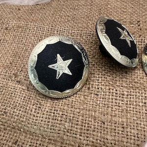 May include: Silver-toned metal star-shaped cabinet knobs with a black center. The knobs have a decorative, etched border and a star in the center. The knobs are shown on a burlap surface.