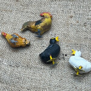 K&ouml;nnte beinhalten: Vier kleine, bemalte Bauernhoftierfiguren: ein Hahn, eine Henne, eine Ente und ein schwarzer Vogel. Der Hahn ist gelb und braun, die Henne ist gelb und orange, die Ente ist wei&szlig; und der schwarze Vogel ist schwarz. Alle haben gelbe Schn&auml;bel und F&uuml;&szlig;e.