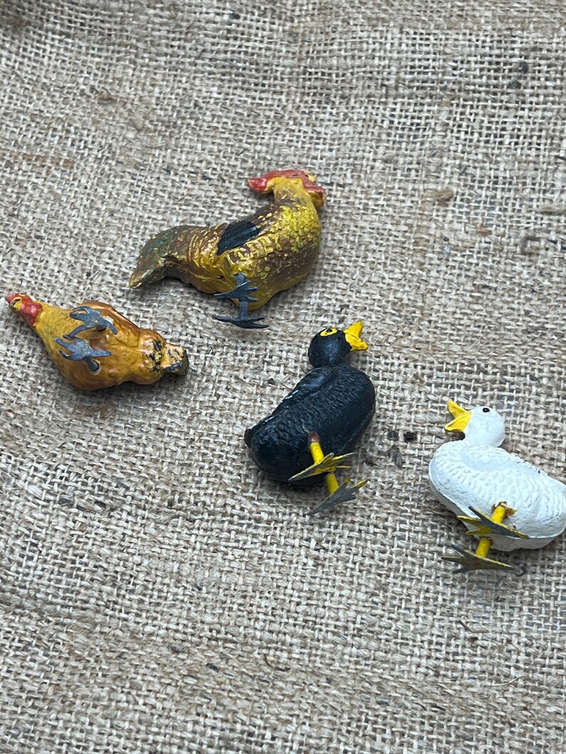 K&ouml;nnte beinhalten: Vier kleine, bemalte Bauernhoftierfiguren: ein Hahn, eine Henne, eine Ente und ein schwarzer Vogel. Der Hahn ist gelb und braun, die Henne ist gelb und orange, die Ente ist wei&szlig; und der schwarze Vogel ist schwarz. Alle haben gelbe Schn&auml;bel und F&uuml;&szlig;e.