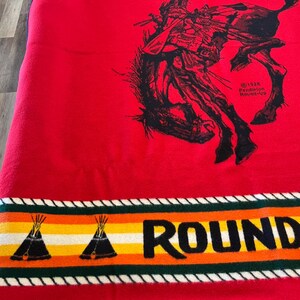 Puede incluir: Manta roja con una ilustraci&oacute;n negra de un vaquero de rodeo montando un bronco. La manta tiene un borde con dise&ntilde;os de tipis y la palabra "ROUND". El borde superior dice "PENDLETON".