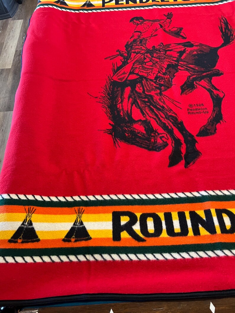 Puede incluir: Manta roja con una ilustraci&oacute;n negra de un vaquero de rodeo montando un bronco. La manta tiene un borde con dise&ntilde;os de tipis y la palabra "ROUND". El borde superior dice "PENDLETON".