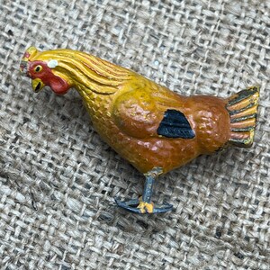 K&ouml;nnte beinhalten: Eine bemalte Metall-H&uuml;hnerfigur. Das Huhn ist haupts&auml;chlich braun mit gelben und orangen Akzenten. Es hat einen roten Kamm und Kehllappen sowie eine schwarze Markierung an der Seite. Die Figur steht auf einer strukturierten Oberfl&auml;che.