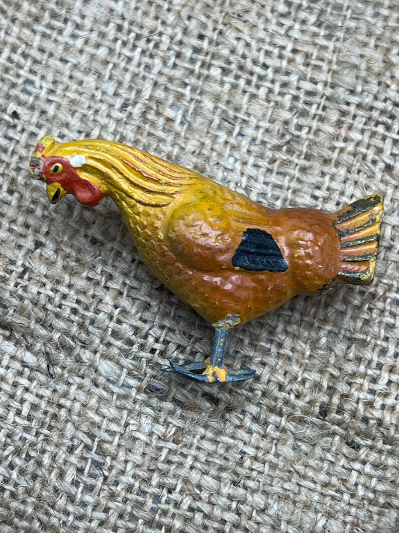 K&ouml;nnte beinhalten: Eine bemalte Metall-H&uuml;hnerfigur. Das Huhn ist haupts&auml;chlich braun mit gelben und orangen Akzenten. Es hat einen roten Kamm und Kehllappen sowie eine schwarze Markierung an der Seite. Die Figur steht auf einer strukturierten Oberfl&auml;che.