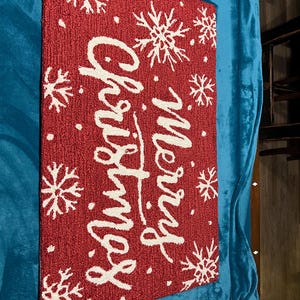 May include: A rectangular red Christmas doormat with white text that reads "Merry Christmas". The mat features white snowflake designs and small white dots. The mat is made of a textured material.