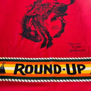 Puede incluir: Manta roja Pendleton con una ilustraci&oacute;n negra de un vaquero montando un caballo salvaje. La manta tiene un borde con las palabras "ROUND-UP" y "PENDLETON" en blanco, amarillo, naranja y verde. Firmada por Wallace Smith.