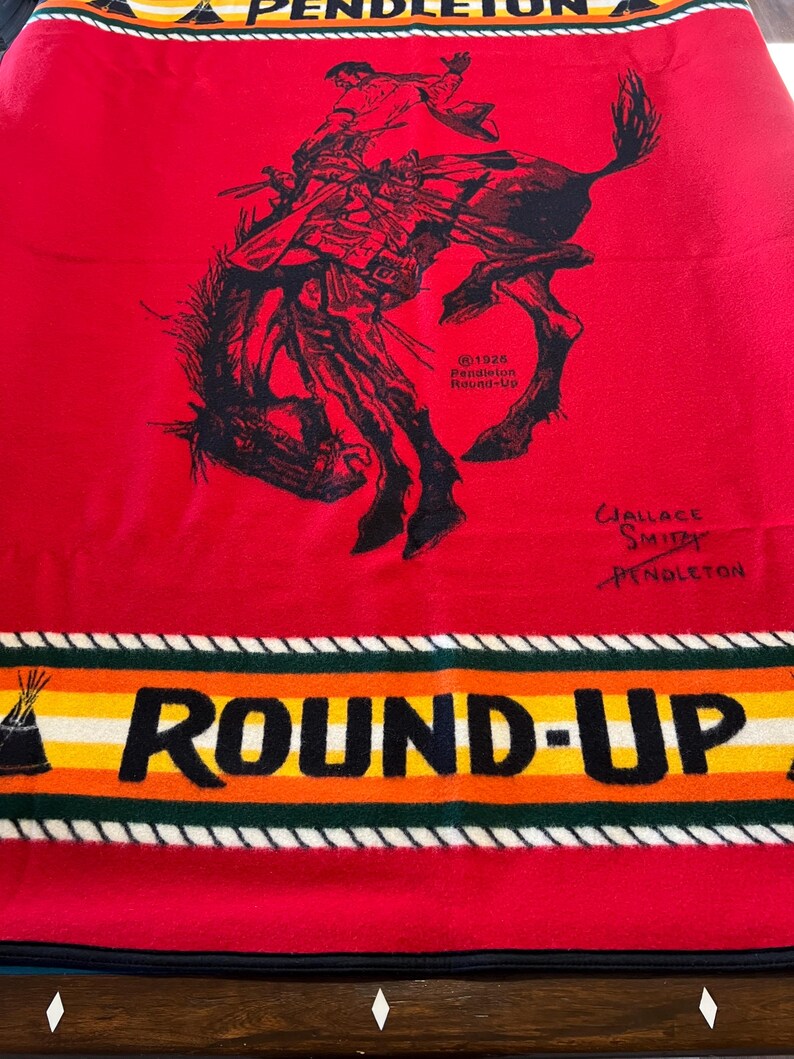 Puede incluir: Manta roja Pendleton con una ilustraci&oacute;n negra de un vaquero montando un caballo salvaje. La manta tiene un borde con las palabras "ROUND-UP" y "PENDLETON" en blanco, amarillo, naranja y verde. Firmada por Wallace Smith.