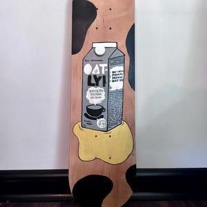 May include: A wooden skateboard deck painted with a black and white cow pattern. The deck features a cartoon illustration of a carton of oat milk with the text "The Original Oat-ly! Barista Edition Oat Drink" and "Totally Vegan. 1L".