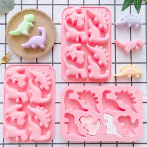 May include: Three rectangular pink silicone molds featuring various dinosaur-shaped cavities. Loose dinosaur-shaped figures in pastel green, purple, pink, and yellow are arranged on a wooden plate and the surrounding surface.