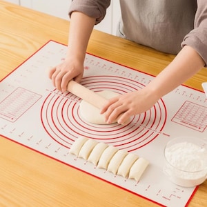 Peut inclure: Un tapis de cuisson rectangulaire en silicone avec des marquages de mesure rouges imprimés, incluant des cercles concentriques et une règle sur les bords. Un rouleau à pâtisserie en bois aplatit la pâte, avec des portions de pâte à côté.