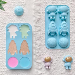 May include: Two light blue silicone molds featuring space-themed cavities including astronauts, rockets, and planets. One mold is filled with various colored substances, while the other is empty. Two small astronaut figurines are placed nearby.
