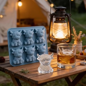 May include: A blue silicone ice cube tray with six Highland cow shapes, a clear ice cow, and a glass with a cow-shaped ice cube. The items are on a wooden table with a lantern and camping tent in the background.