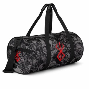 May include: A cylindrical duffel bag in black and grey with a repeating abstract design. It has red graphic symbols and black handles and a shoulder strap. The bag is designed for carrying items.