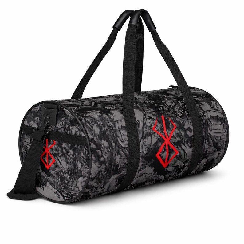 May include: A cylindrical duffel bag in black and grey with a repeating abstract design. It has red graphic symbols and black handles and a shoulder strap. The bag is designed for carrying items.