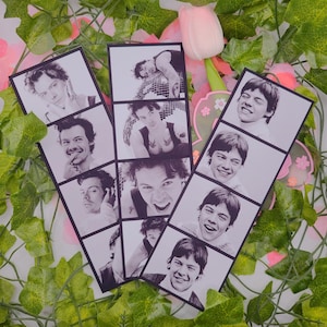 May include: Black and white photo strips featuring multiple images of a person. The strips are arranged on a surface with green vines, pink flowers, and a pink tulip. The photos show the person in various poses.