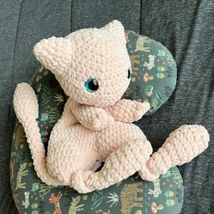 Puede incluir: Un juguete de peluche rosa de crochet con forma de gato, con ojos azules, sentado en una almohada redonda con estampado de bosque.