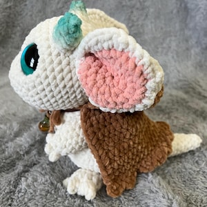 May include: A white crocheted dragon plush toy with a pink and brown crocheted cape. The dragon has large blue eyes and a green and blue horn.