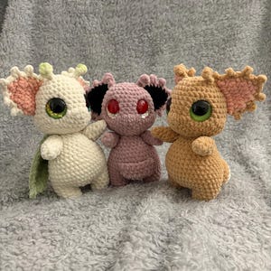 Może przedstawiać: Trzy pluszowe zabawki wykonane na szydełku, jedna biała z zielonymi akcentami, jedna różowa z czarnymi akcentami i jedna brązowa z różowymi akcentami. Każda zabawka ma duże, kolorowe oczy.