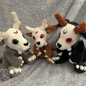 May include: Three handmade crochet plush toys in a row. Each has a skull-like head with red eyes and a crescent moon. The plush toys are in shades of gray, brown, and black, with white and brown accents. They have horns and are sitting.