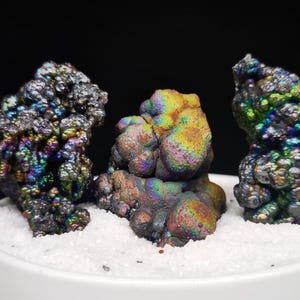 Könnte beinhalten: Drei irisierende Mineralproben mit dunkler, strukturierter Oberfläche. Die Mineralien zeigen eine Reihe von Farben, darunter Blau, Grün, Gelb und Violett. Sie sind auf einem Bett aus weißen Granulaten in einem weißen Keramikbehälter angeordnet.