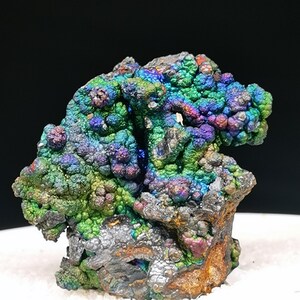 Könnte beinhalten: Ein Mineralexemplar mit botryoidaler Struktur und einer metallisch schillernden Oberfläche. Die Ansammlung zeigt lebhafte Grün-, Blau-, Violett- und Goldtöne mit einer rauen, texturierten Basis, die braune und graue Mineralablagerungen aufweist.