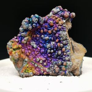 Könnte beinhalten: Ein farbenfrohes Mineralpräparat mit strukturierter Oberfläche. Der Stein zeigt eine lebendige Farbpalette, darunter Lila, Blau, Orange und Silber. Das Mineral befindet sich auf einer weißen Oberfläche und einem schwarzen Hintergrund.