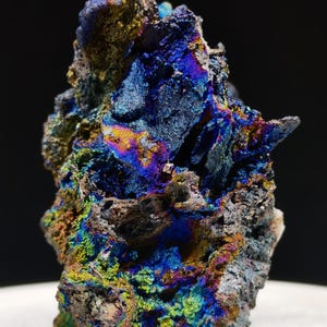 Könnte beinhalten: Nahaufnahme einer Mineralprobe mit rauer, strukturierter Oberfläche. Das Mineral zeigt eine lebendige Farbpalette, darunter Blau-, Grün-, Gelb- und Violetttöne, wodurch ein schimmernder, irisierender Effekt entsteht. Die Gesamtform ist unregelmäßig und gezackt.