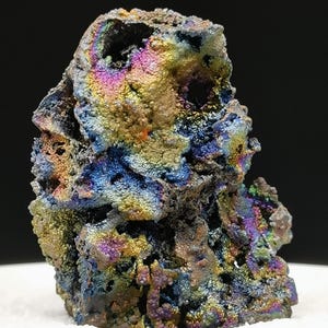 Puede incluir: Primer plano de un espécimen mineral con una superficie rugosa y texturizada. El mineral muestra una vibrante gama de colores, incluyendo azules iridiscentes, púrpuras, verdes y dorados. El espécimen se encuentra sobre una superficie blanca contra un fondo negro.