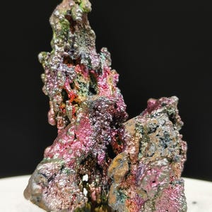 Puede incluir: Primer plano de un espécimen mineral con una superficie texturizada e iridiscente. Los colores van del púrpura intenso y rosa al verde y dorado. El mineral tiene una estructura rugosa y cristalina y se asienta sobre una superficie blanca.