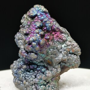 Puede incluir: Primer plano de un espécimen mineral con una superficie texturizada y burbujeante. El mineral muestra una gama de colores, incluyendo azules, morados y toques de rosa y verde. El mineral se encuentra sobre una superficie blanca sobre un fondo negro.