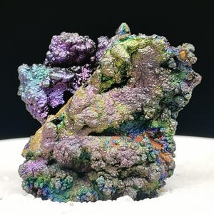 Könnte beinhalten: Nahaufnahme einer Mineralprobe mit strukturierter Oberfläche. Das Mineral zeigt eine lebendige Farbpalette, darunter Lila, Grün, Blau und Orange. Die Probe befindet sich auf einer weißen Oberfläche und einem schwarzen Hintergrund.
