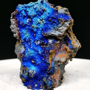 Könnte beinhalten: Ein rohes Mineralexemplar mit einer leuchtend irisierenden, blauen Metalloberfläche und strukturierten, zerklüfteten Formationen. Der Stein zeigt Flecken in tiefem Kobaltblau, durchsetzt mit Bereichen aus oxidierten orangefarbenen, braunen und dunkelgrauen Mineralablagerungen.