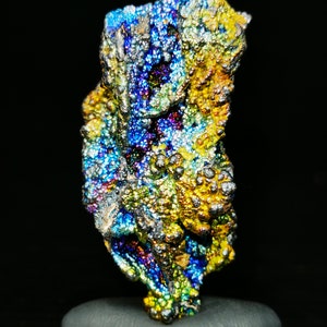 Könnte beinhalten: Ein regenbogenfarbener, rauer Mineralienproben mit einer unebenen, strukturierten Oberfläche. Das Mineral wird auf einem grauen Stein präsentiert.