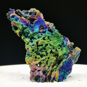 Könnte beinhalten: Nahaufnahme einer farbenfrohen Mineralprobe mit rauer, strukturierter Oberfläche. Das Mineral zeigt eine lebendige Farbpalette, darunter Blau-, Grün-, Lila- und Goldtöne. Die Probe ist vor einem weißen Hintergrund platziert.