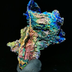 Könnte beinhalten: Ein irisierendes blaues, grünes und violettes Mineralstück mit einer rauen, strukturierten Oberfläche. Das Mineral wird auf einem kleinen, grauen Sockel präsentiert.