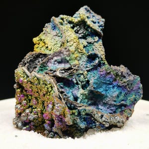 Könnte beinhalten: Nahaufnahme einer Mineralprobe mit rauer, strukturierter Oberfläche. Der Stein zeigt eine lebendige Farbpalette, darunter Blau-, Grün-, Gelb- und Lilatöne. Das Mineral liegt auf einer weißen Oberfläche vor einem schwarzen Hintergrund.