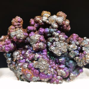 Könnte beinhalten: Nahaufnahme eines Mineralexemplars mit einer Ansammlung von abgerundeten Formationen. Das Mineral weist einen metallischen Glanz mit Farben von Lila und Silber bis Bronze und Gold auf. Das Exemplar ist vor einem schwarzen Hintergrund platziert.
