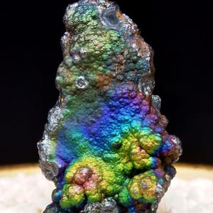 Puede incluir: Un espécimen mineral iridiscente, multicolor, rugoso y abultado. El mineral tiene una base gris oscura con un arcoíris de colores, incluyendo verde, azul, morado y rojo.