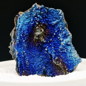 Könnte beinhalten: Nahaufnahme einer leuchtend blauen Mineralprobe mit strukturierter Oberfläche. Das Mineral weist eine Mischung aus Dunkelblau, Hellblau und Andeutungen von Lila und Braun auf. Kleine, silbernfarbene kristalline Strukturen sind über die Oberfläche verstreut.